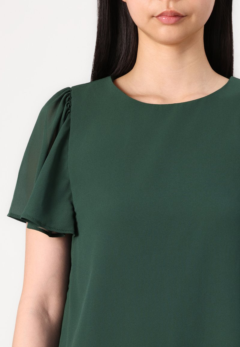 Blouse verte avec un col rond, des manches courtes volantées et une texture lisse et mate. Le tissu semble léger et fluide.