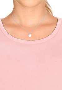 Silberne Kette mit einer dünnen, gedrehten Kette und einem glatten, runden Anhänger, getragen über einem hellrosa, gerippten Shirt.