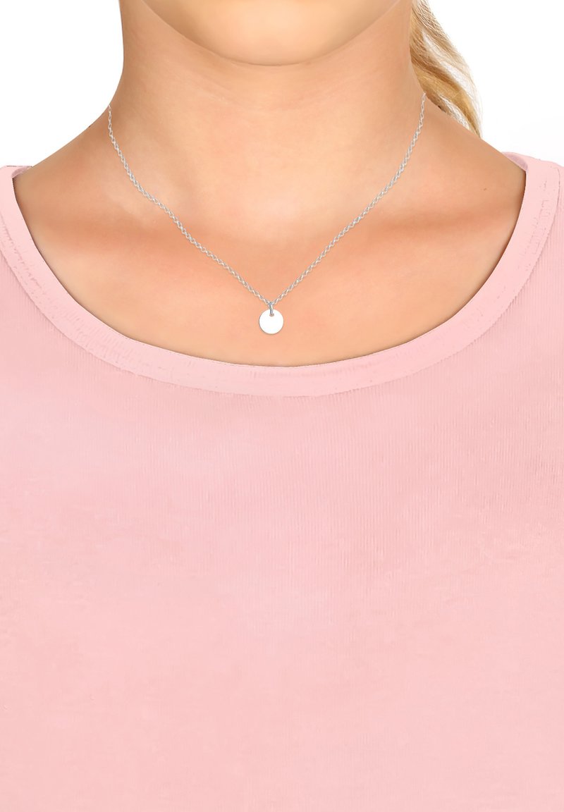 Silberne Kette mit einer dünnen, gedrehten Kette und einem glatten, runden Anhänger, getragen über einem hellrosa, gerippten Shirt.
