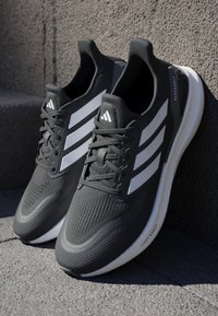Baskets de sport noires avec surface texturée, rayures blanches et lacets. Dotées d'une semelle rembourrée étiquetée "LIGHTBOOST" pour le confort.