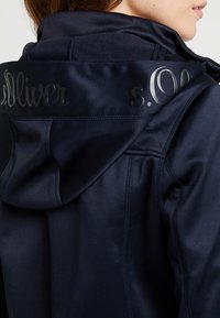Marinblå huva jacka med en slät yta, prydd med en blank svart "s.Oliver"-logotyp på ryggen och kontrasterande sömdetaljer.