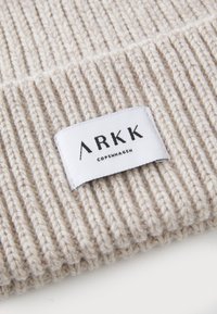 Beige ribbestrikket stoff med et vevd merke som har "ARKK COPENHAGEN" i svart tekst. Teksturen er myk og litt elastisk.