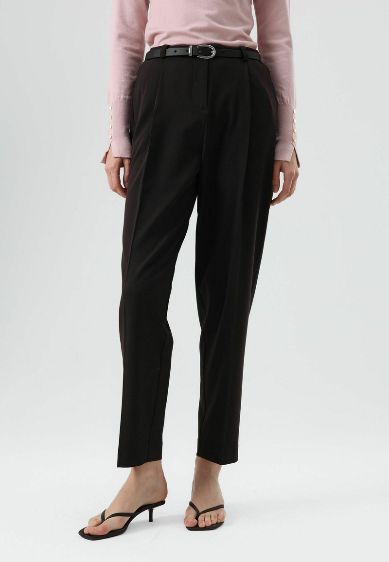 Pantalon noir sur mesure à taille haute, coupe droite et plis marqués, associé à un pull rose pâle avec des accents de boutons sur les manches.