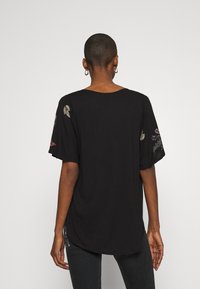 Svart topp med korta ärmar och rundad halsringning, med blommönster broderade på ärmarna. Mjukt tyg med en avslappnad passform och en något krökt nederkant.