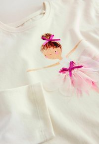 Cremefarbener Stoff mit einer gedruckten und genähten Ballerina, die einen rosa Tüllrock und eine Haarschleife trägt, mit detaillierter Stickerei am Ärmel.