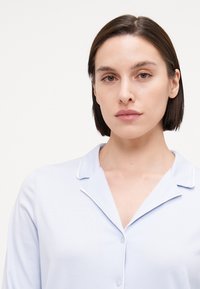 Ljusblå långärmad pyjamasöverdel med snedkrage, knappstängning och vit piping längs kragen och knappslån. Slät textur.