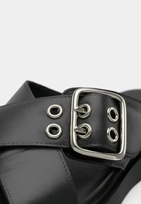Bride de sandale en cuir noir avec une grande boucle rectangulaire argentée et quatre œillets métalliques sur un fond uni.