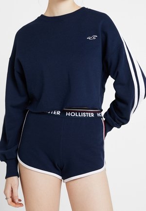 Persona con sudadera corta de manga larga azul marino con rayas blancas en las mangas y pantalones cortos a juego en azul marino con ribete blanco y cintura con el logo de Hollister.