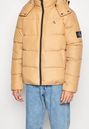 Homme portant une doudoune beige avec une fermeture éclair noire, logos Calvin Klein sur la poitrine et la manche, associée à un jean bleu clair et des bagues argentées.