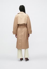 Μπεζ παλτό τύπου trench με ζώνη στη μέση, διπλό κούμπωμα εμπρός και φαρδιές πέτο. Διαθέτει μακριά μανίκια και ελαφρώς κοντό πίσω μέρος.