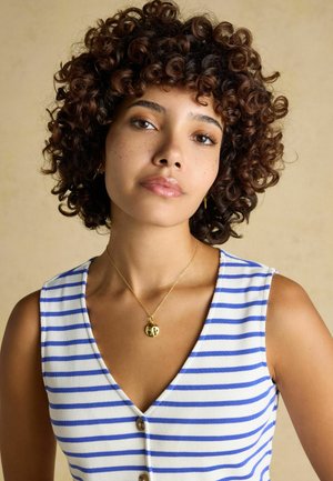 Junge Frau mit lockigem braunem Haar, die ein ärmelloses Top mit blau-weißen Streifen und eine goldene Anhänger-Halskette vor einem beigefarbenen Hintergrund trägt.