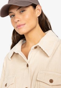 Veste en velours côtelé beige avec un col texturé, deux poches avant et des boutons marron. Le modèle porte également une casquette sombre.