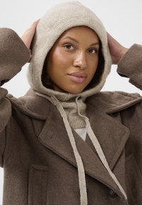 Barna gyapjú peacoat széles hajtókákkal, bézs kötött kapucnival és húzózsinórokkal. A textúra puha, lazán illeszkedik, és elülső zsebei vannak.