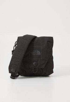Kompakt sort crossbody-taske med klaplukning, justerbar rem og The North Face-logo foran mod en ensartet lys baggrund.
