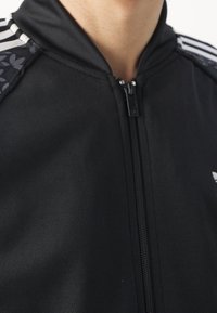 Černá bunda na zip z hladké, elastické látky. Má proužkové detaily na ramenou a výrazný květinový vzor na jedné straně.