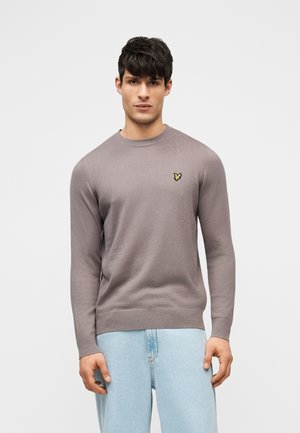 Jeune homme portant un pull ras du cou taupe avec un petit logo d'oiseau jaune sur la poitrine et un jean bleu clair, sur fond uni.