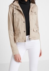 Beige knäppt och dragkedjade jacka med fotor fickor, bärs över en svart polotröja och vita åtsittande byxor.