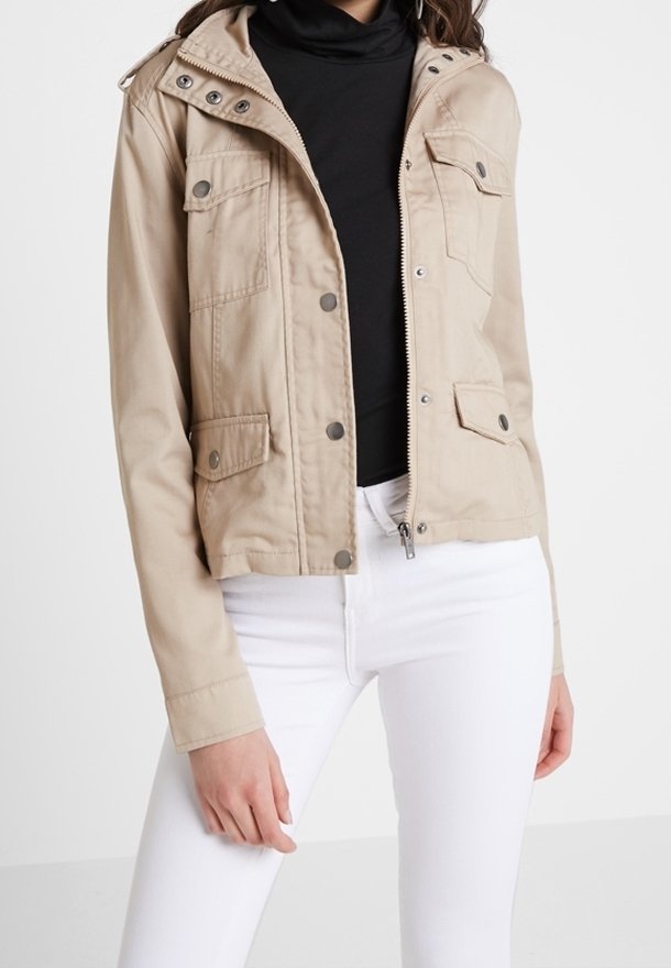 Beige knäppt och dragkedjade jacka med fotor fickor, bärs över en svart polotröja och vita åtsittande byxor.