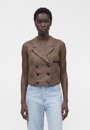 Donna che indossa un gilet marrone senza maniche doppiopetto con grandi bottoni abbinato a jeans azzurri a vita alta, in piedi davanti a uno sfondo bianco.