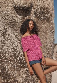 Lyserød off-shoulder top med blomsterprint, indsnøret halsudskæring og løse ærmer, kombineret med denimshorts foran en klippefyldt baggrund.
