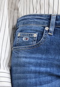 Los vaqueros de mezclilla azul presentan un diseño clásico de cinco bolsillos, remaches de cobre y un pequeño logo bordado en el bolsillo trasero. Textura suave.