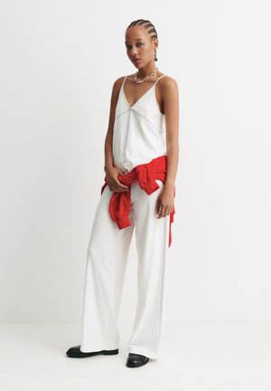 Top bianco senza maniche con scollatura a V, abbinato a pantaloni bianchi a gamba larga. Giacca rossa legata in vita. Scarpe nere basse. Collana in argento a catena.