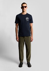 Marineblå bomulds-T-shirt med korte ærmer, frontgrafik af en løve og tekstdetalje. Samlet med olivengrønne bukser og sorte snørebåndsstøvler.
