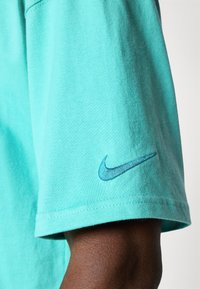 Camiseta de algodón turquesa con manga corta y dobladillo cosido, que presenta un logo de Nike en azul oscuro en la manga. Textura suave.