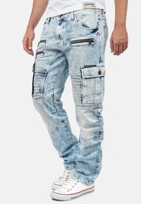 Light blue distressed denim jeans with multiple zippered pockets, ribbed accents, and a relaxed fit, paired with white sneakers.