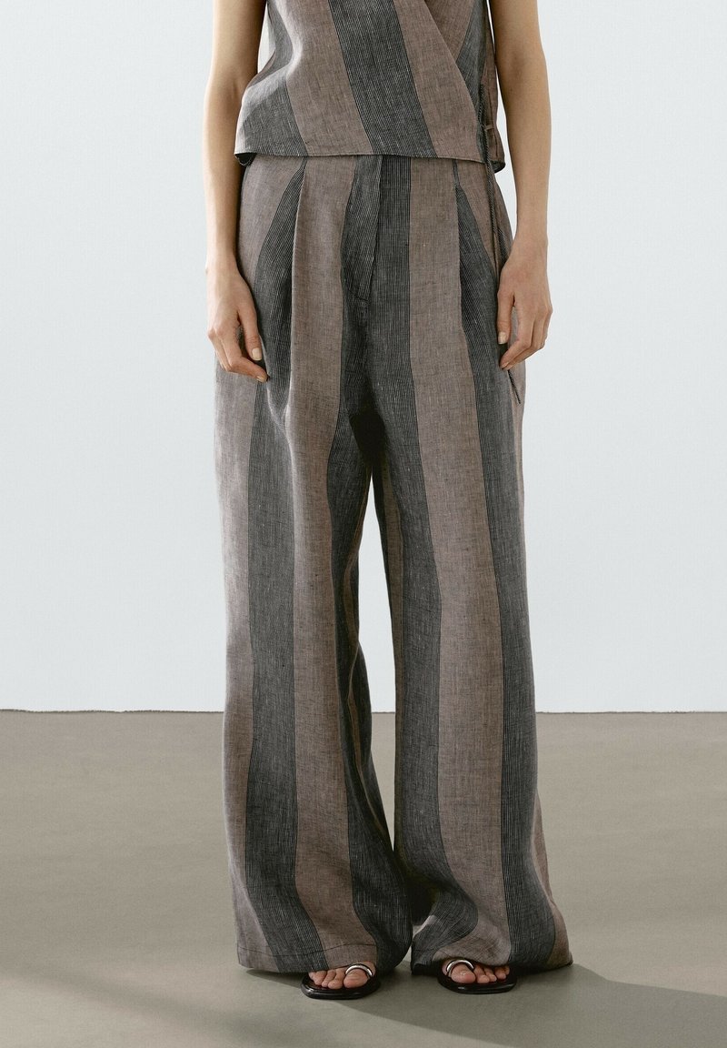 Femme portant un pantalon large et ample à jambes larges avec des rayures verticales grises et beiges, associé à un haut rayé assorti et des sandales noires.