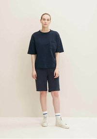 Navy blue oversized t-shirt with a chest pocket, paired with navy blue knee-length shorts. Model wears white sneakers with striped socks.