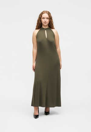 Femeie cu păr roșcat și creț, purtând o rochie maxi verde măsliniu fără mâneci și pantofi cu toc negri, stând în picioare pe un fundal alb simplu.