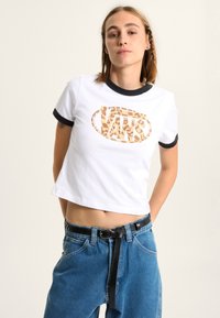 Short-sleeve white crop top with black ribbed collar featuring a tan leopard print "VANS" logo at the center. Paired with high-waisted blue jeans.