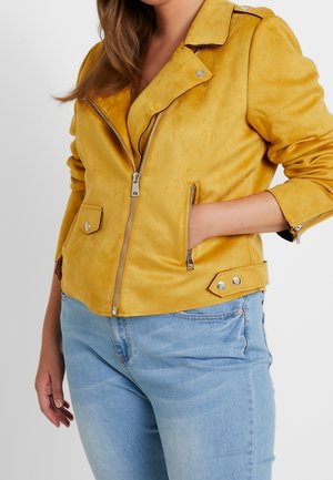 Femme portant une veste de motard en daim jaune avec des fermetures éclair argentées et un jean bleu clair, une main dans la poche et l'autre détendue le long du corps.