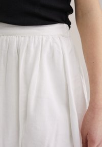 White linen skirt with a high waistband, featuring subtle pleats. Close-up of fabric texture and smooth finish, with a hand visible at the side.