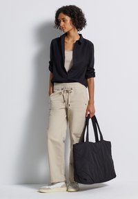 Femme portant une chemise noire à boutons sur un pantalon beige et des baskets blanches, tenant un grand sac fourre-tout noir matelassé, debout contre un mur blanc.