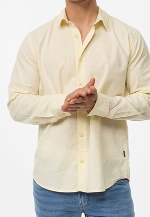 Mann trägt ein blassgelbes Hemd mit Knöpfen und blaue Jeans, die Hände vor dem Körper verschränkt, vor einem schlichten weißen Hintergrund.