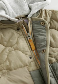 Gequiltte jas met een mix van beige en olijfgroene materialen. Beschikt over een grijze capuchon, oranje rits en knoopdetails. Soepele textuur.