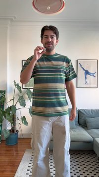 Grün gestreiftes T-Shirt mit horizontalen Streifen in Grün-, Gelb- und Blautönen. Getragen zu hellen Cargohosen; Innenraumszenerie.