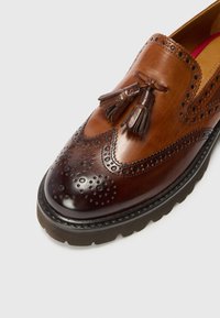 Brūna ādas slip-on kurpe ar brogue detaļām, ar pušķu akcentiem un teksturētu zoli. Gluda un pulēta virsma.
