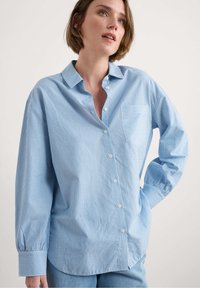 Gaiši zils krekls ar baltām vertikālām svītrām, krūšu kabata un garām piedurknēm ar elastīgām aprocēm. Izgatavots no gludas auduma.