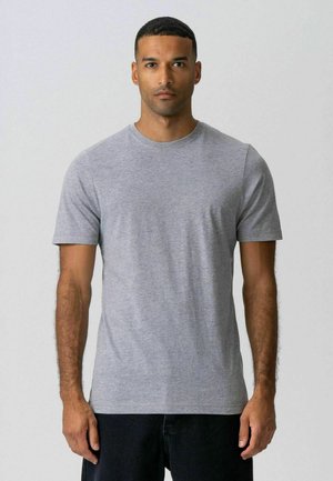 Mand stående fremadvendt iført en ensfarvet grå T-shirt med korte ærmer og mørke bukser mod en lys baggrund.