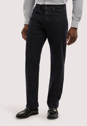 Mann trägt schwarze Jeans mit geradem Bein, schwarze Leder-Loafer und ein hellgraues, langärmliges Hemd mit Knöpfen vor neutralem Hintergrund.