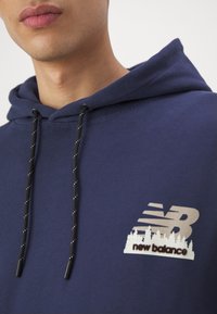 Gros plan d'une personne portant un sweat à capuche bleu marine New Balance avec des cordons noirs et un logo avec des arbres sur la poitrine.