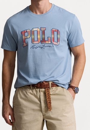 T-shirt en coton bleu clair avec un motif à carreaux multicolores "POLO" en graphisme et logo en écriture. Associé à un short beige et une ceinture tressée marron.