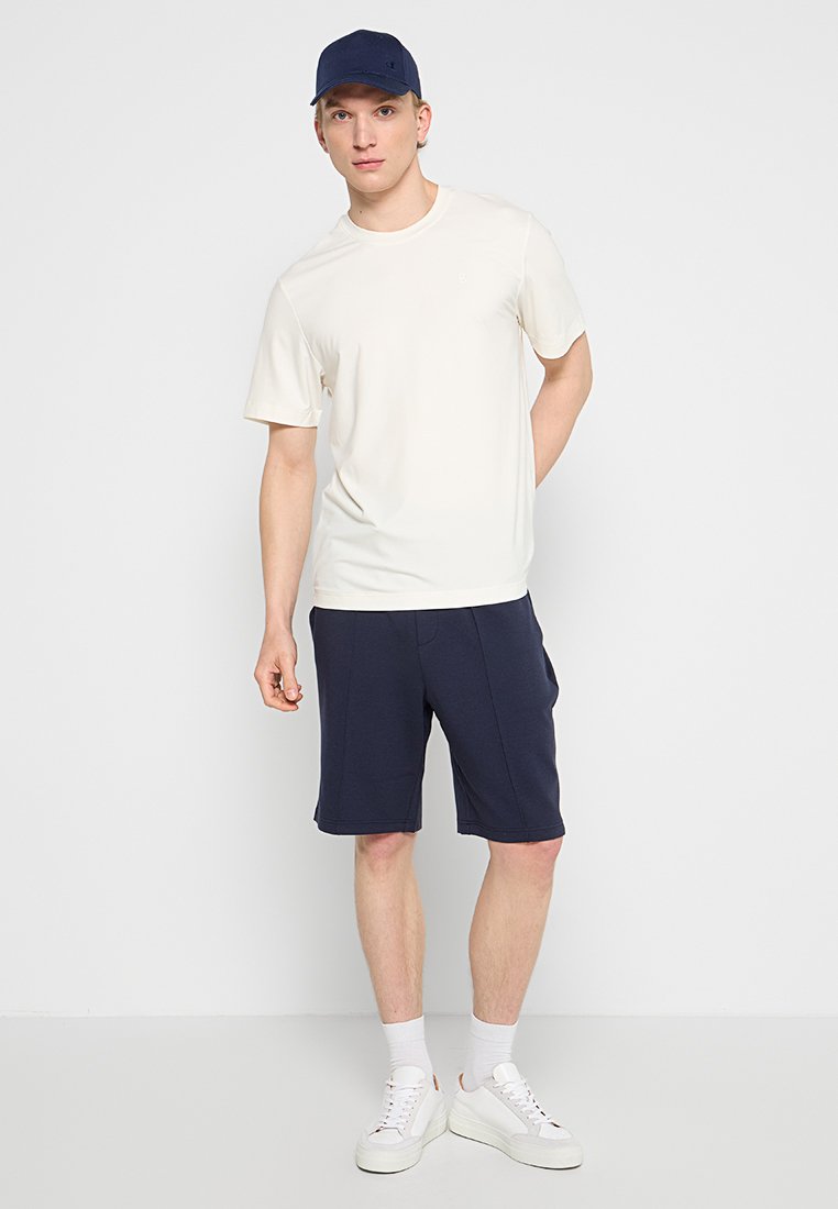Hellbeige T-Shirt mit kurzen Ärmeln und marineblauen Shorts, kombiniert mit einer marineblauen Mütze. Weiße Sneakers mit strukturierten Sohlen. Schlichte Gestaltung, keine sichtbaren Muster.