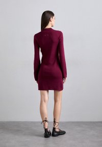 Egy nő áll, háttal nézve, testhezálló, hosszú ujjú bordó ruhát és fekete fűzős lapos cipőt viselve, egy sima szürke háttér előtt.