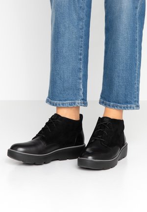 Bottines à lacets en cuir noir et daim portées avec un jean bleu retroussé sur une surface blanche.