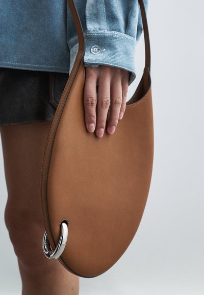 Hnedá kožená kabelka s oblým oválnym tvarom, doplnená strieborným krúžkom a minimalistickými detailmi šitia. Ruka drží kabelku.