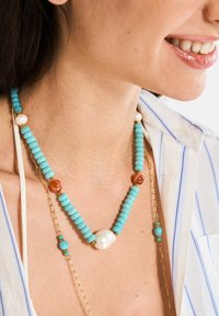 Collana di perle turchesi con accenti in catena dorata, una grande perla bianca e piccole perle arancioni e verdi, presentata in un design a strati.